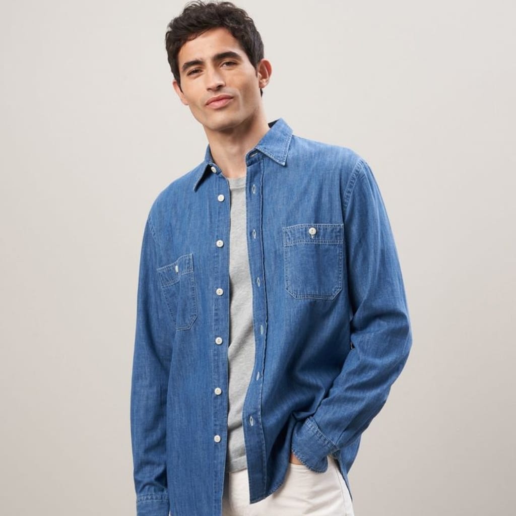 A young man wearing a light blue chambray shirt layered over a gray t-shirt, with his left hand in his pocket, standing against a neutral background.