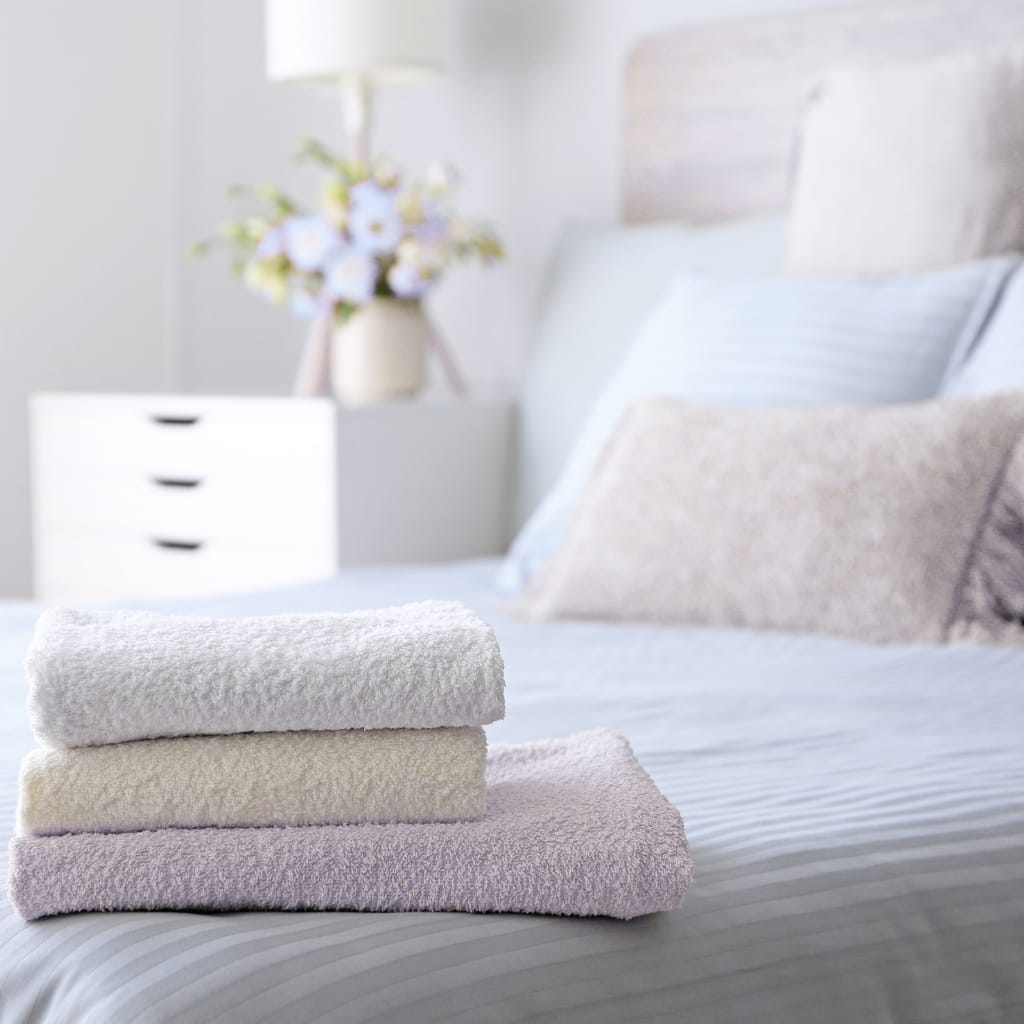 A neatly folded stack of three towels on a bed, with a bedroom in the background featuring a bedside table, a vase with flowers, and pillows.
