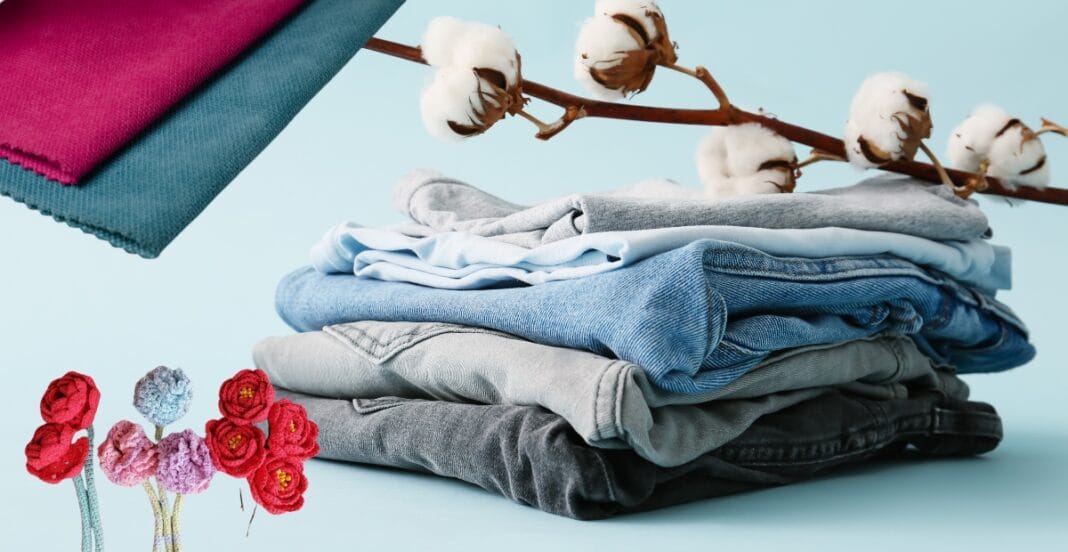 A neatly folded stack of jeans and casual clothing in shades of blue, gray, and black, with a cotton branch and some colorful fabric swatches in the background, and small embroidered flower artwork in the foreground.