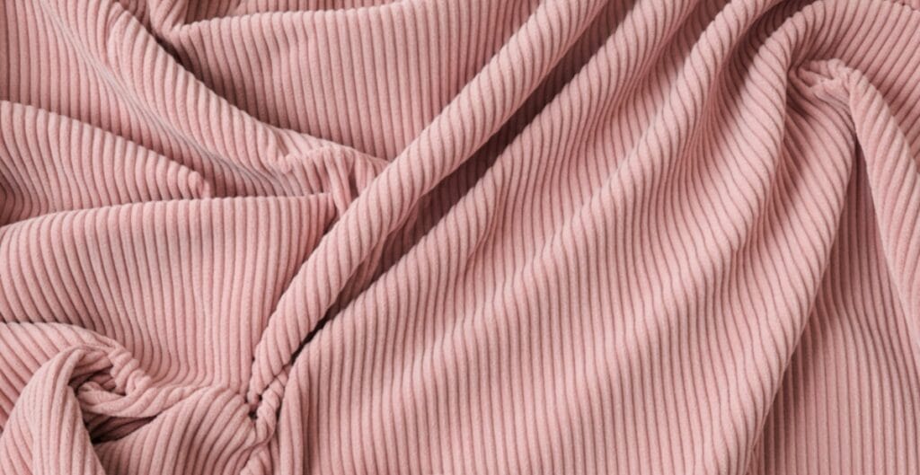 Close-up image of soft, pink ribbed corduroy fabric with visible vertical ridges and folds.