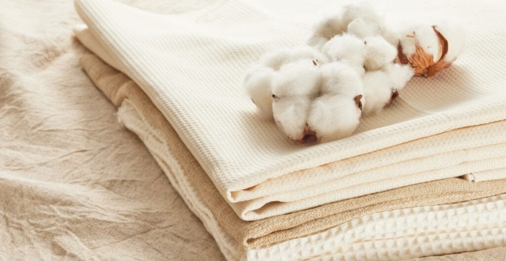 A stack of folded beige and cream-colored fabrics with a small cluster of cotton boll on top, placed on a textured fabric surface.