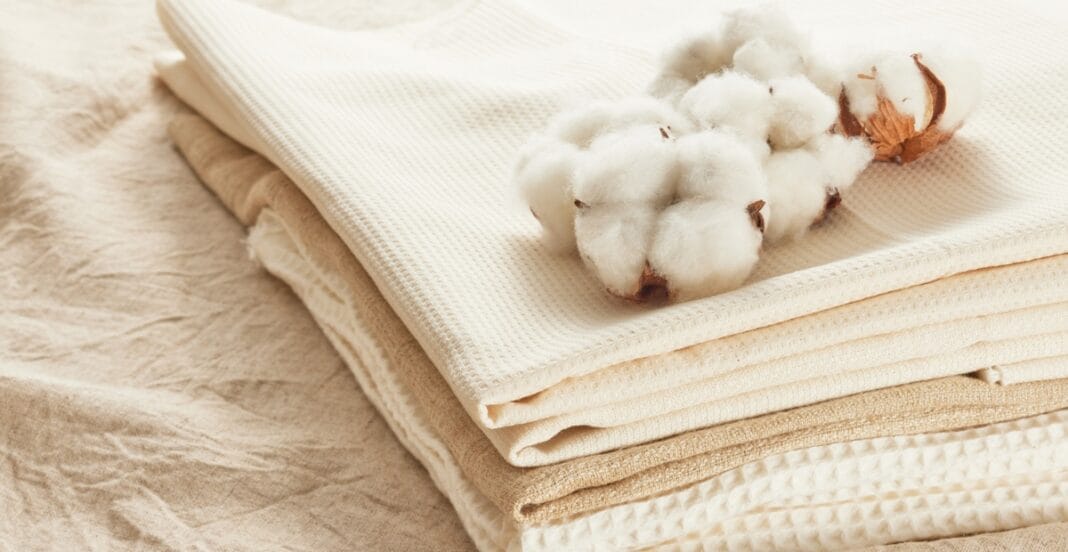 A stack of folded beige and cream-colored fabrics with a small cluster of cotton boll on top, placed on a textured fabric surface.