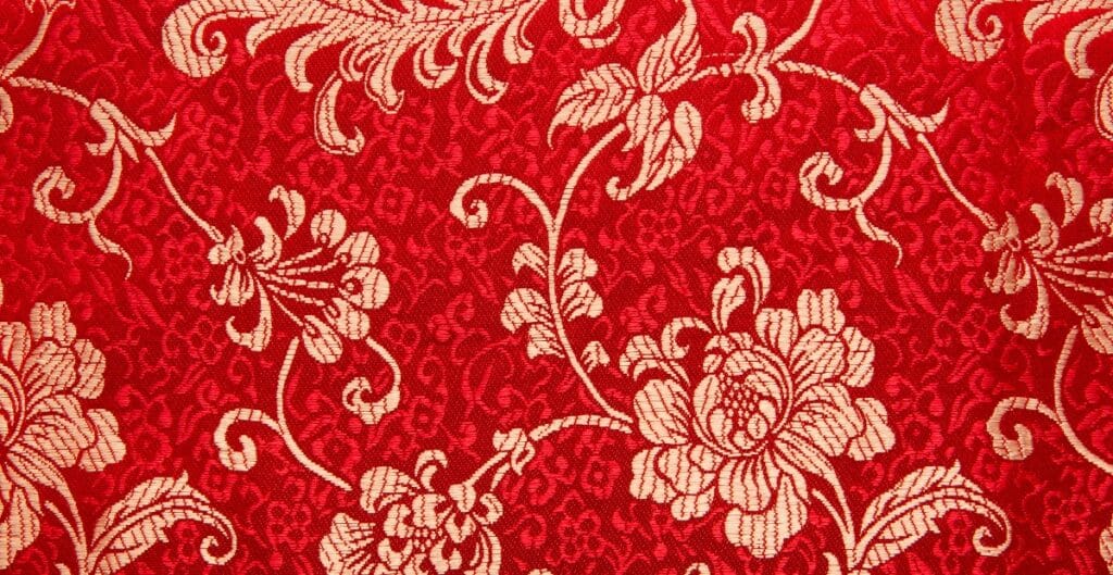 Close-up image of a red designer cotton fabric with an intricate cream floral pattern.