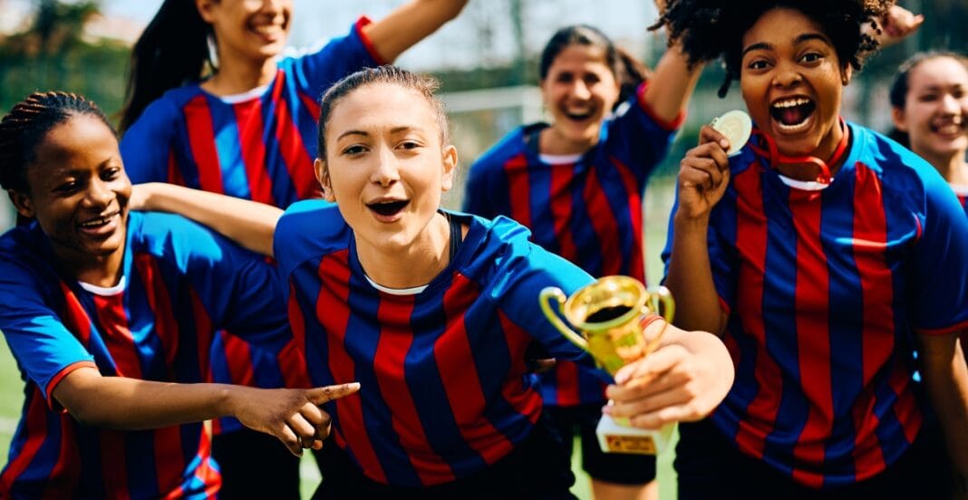 A group of female soccer players wearing blue and red striped jerseys celebrating on the field. One woman at the front is holding a small trophy, and another is pointing at her. They are smiling, cheering, and showing enthusiasm.