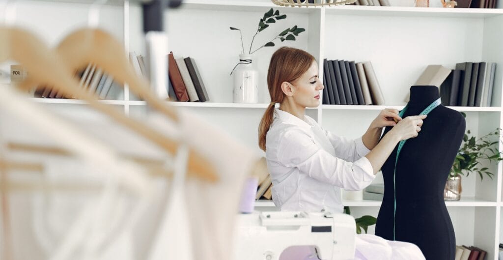 A woman measuring a dress shirt fabric on a mannequin in a well-organized workspace with shelves of books and plants in the background.