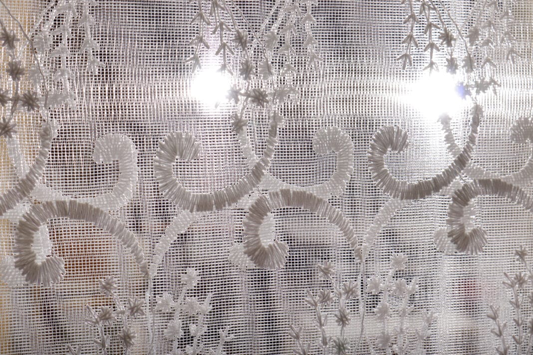 A close-up of a sheer lace curtain with intricate embroidered floral and swirling patterns, allowing light to pass through.