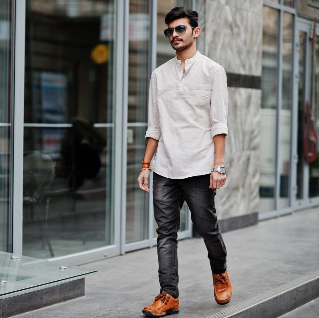A man walking outdoors wearing a light-colored linen shirt, dark jeans, brown boots, sunglasses, and accessories.