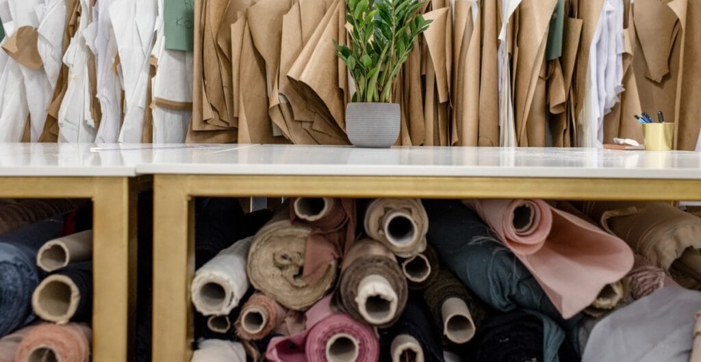 A storage area with a table and fabric rolls underneath it. The table has a potted plant, a mug with pens, and some paper clips. Behind the table, there are neatly arranged brown and white fabric bundles hanging vertically, and below, rolls of fabric in various colors are stored.