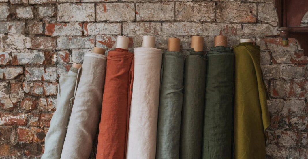 Several rolls of fabric in neutral and earthy tones hanging against a rustic brick wall.