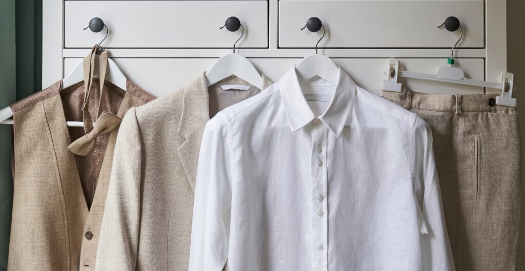 A close-up of a wardrobe with four hanging garments: a beige blazer with a bow tie, a beige suit jacket, a white button-up shirt, and beige trousers, hanging on white and black hooks.