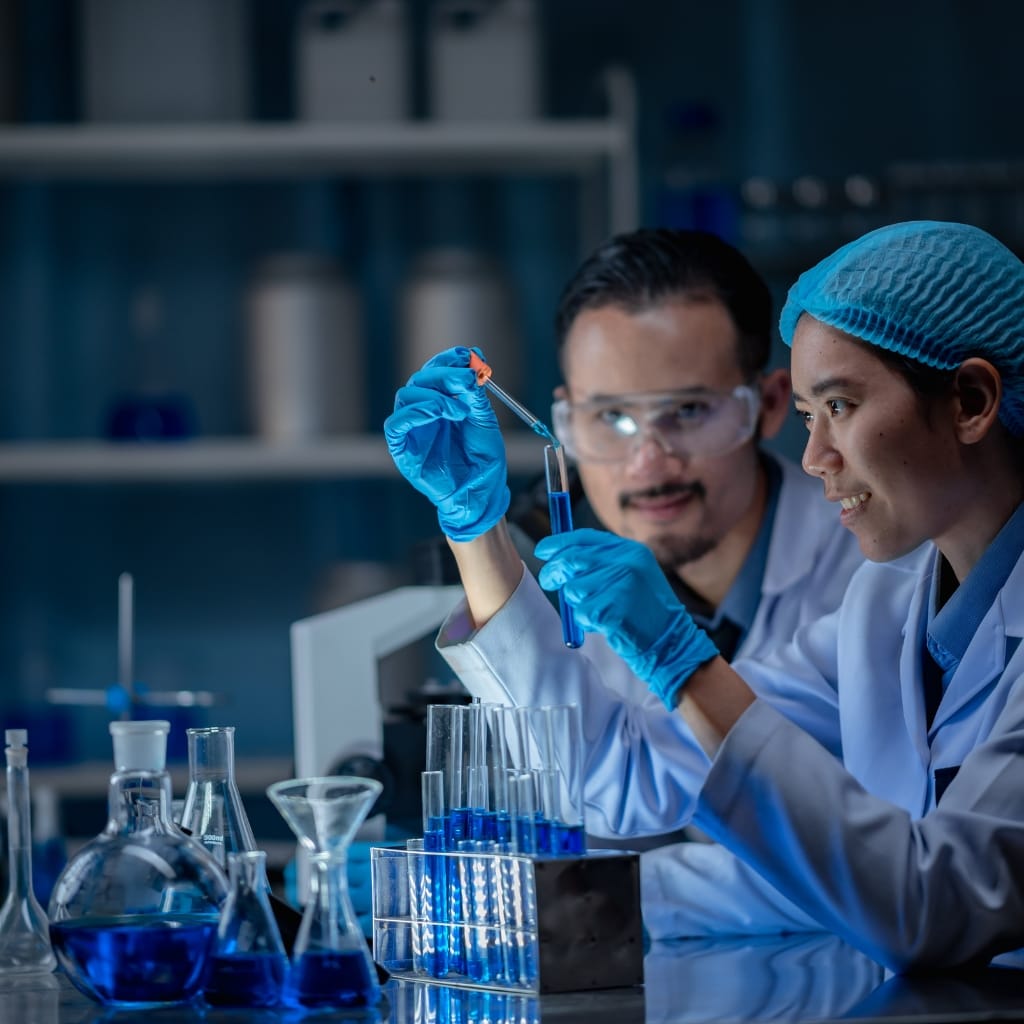 Two scientists working in a laboratory, using pipettes and test tubes filled with a blue liquid, with scientific glassware and equipment around them.
