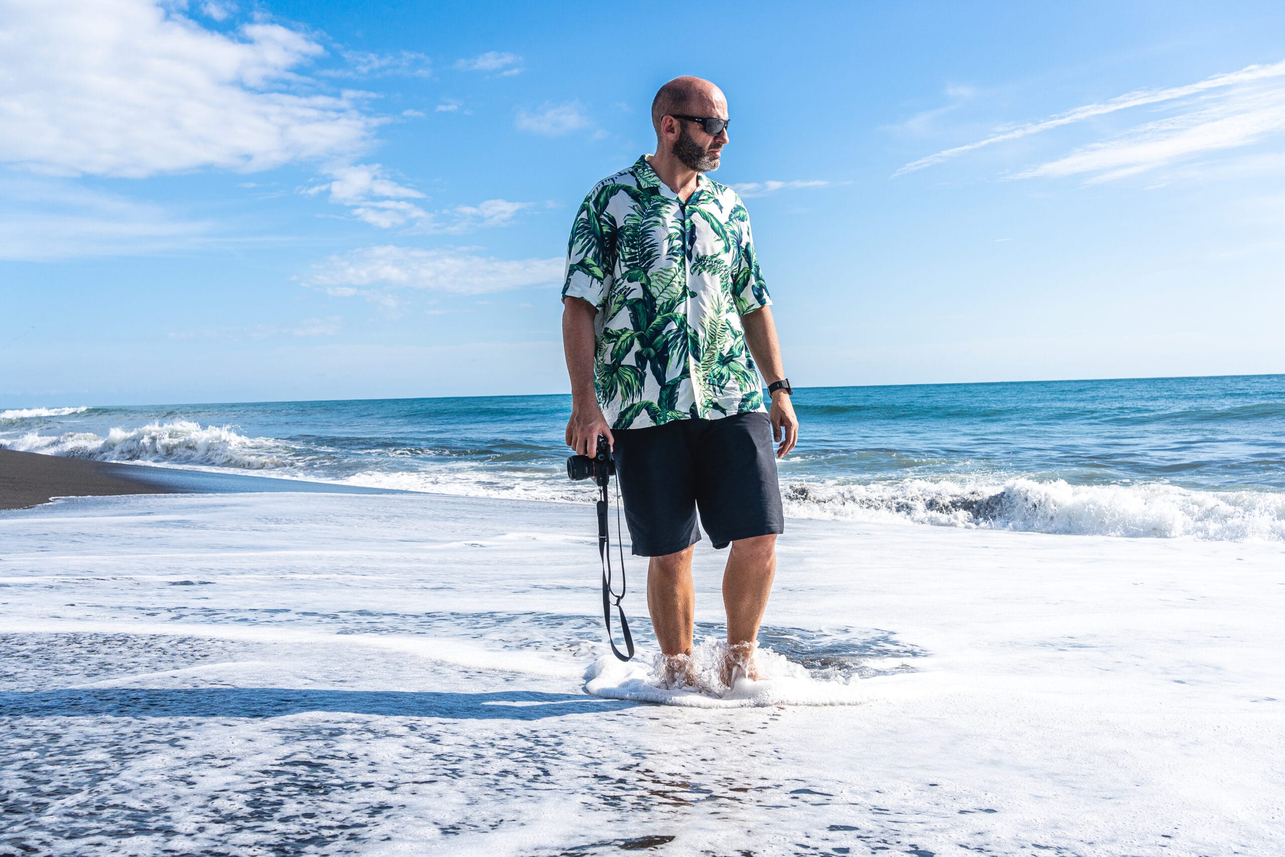 A man standing in the surf at the beach, wearing a vibrant tropical men's beach shirt and sunglasses, holding a camera, with a clear blue sky and ocean in the background.