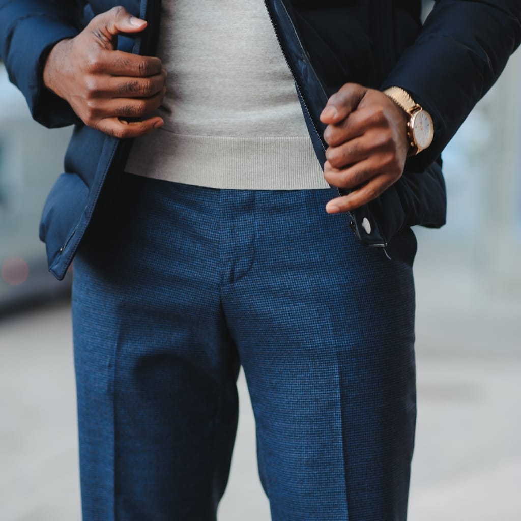 Close-up of a person wearing chambray blue pants, with hands adjusting a dark jacket over a light grey sweater, and a gold wristwatch on their left wrist.