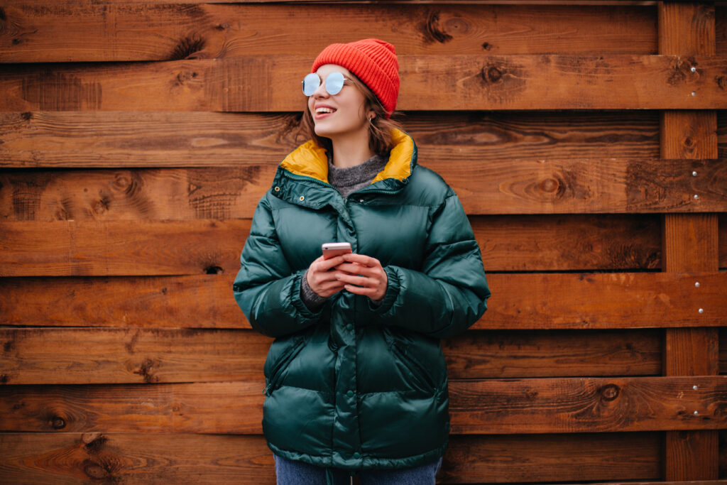 A woman wearing a teal Columbia insulated coat with a yellow lining, standing against a wooden fence, holding a phone, and smiling with sunglasses and a red knit hat.