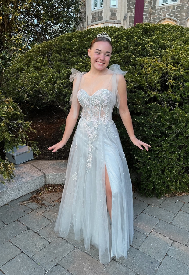 A young woman in a light coral prom dress with floral embellishments, standing outdoors on a paved area with greenery and a stone building in the background, smiling and posing.