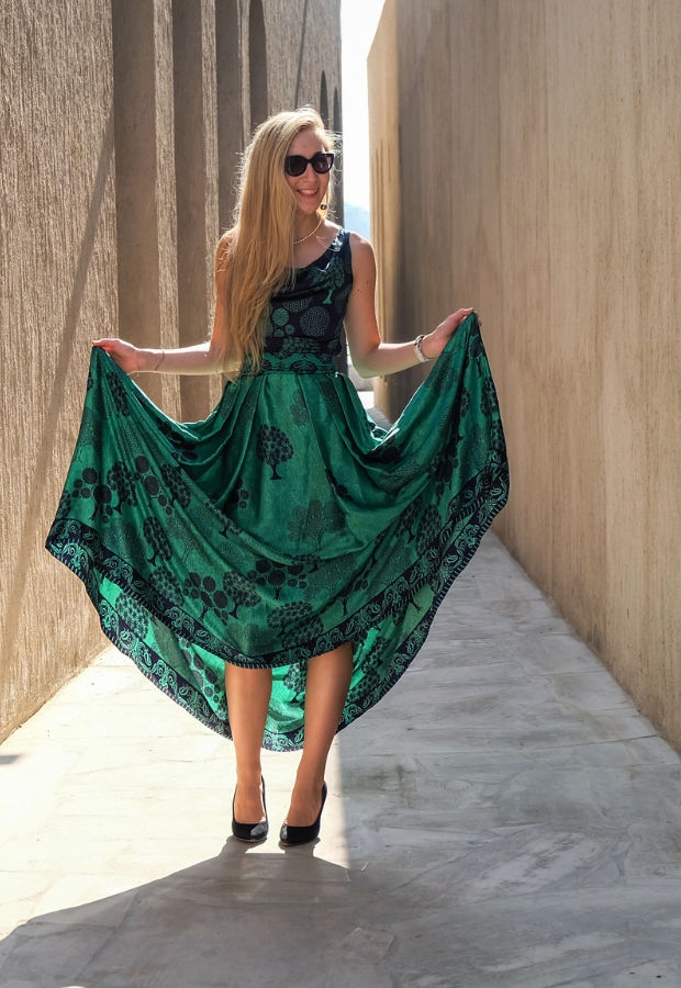 A woman in a vibrant emerald green dress standing in a narrow alleyway, holding out the sides of her full, flowing skirt with black high heels and wearing sunglasses.