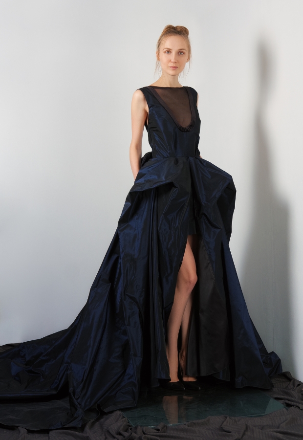 A young woman wearing a striking midnight blue prom dress with a high slit, standing against a plain background. The dress features a sheer black neckline and an elaborate, voluminous skirt with a train.
