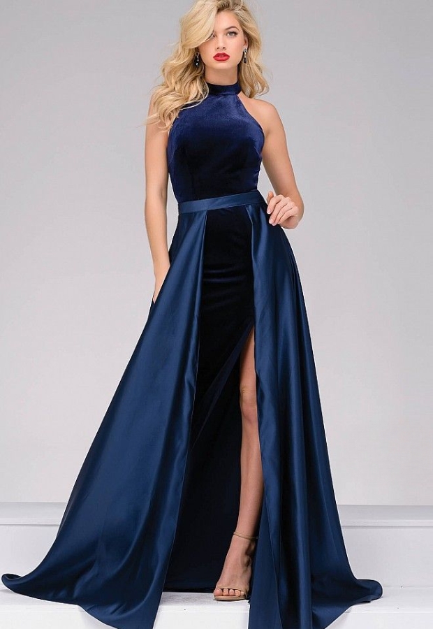 Elegant woman wearing a navy velvet prom dress with a high neckline, fitted bodice, and a dramatic high slit in the skirt. The dress features a shiny, satin-like overlay that flows to the floor, creating a regal and luxurious look. She has long, wavy blonde hair and wears nude heels.