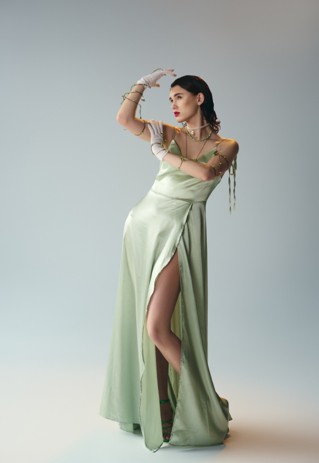 A woman wearing an elegant, light green satin prom dress with a high slit, accessorized with jewelry and gloves, posing against a plain background.