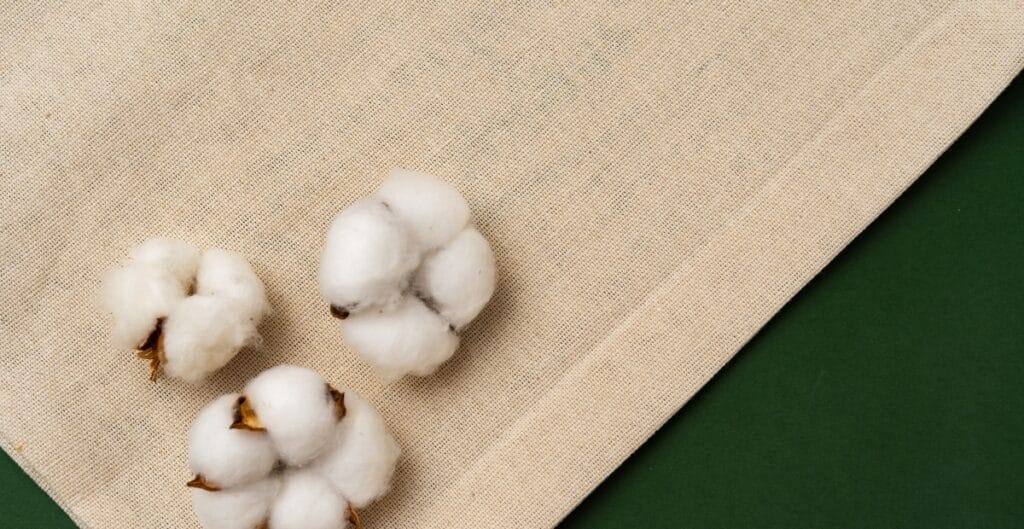 Close-up image of raw cotton bolls placed on a beige fabric surface with green fabric in the background.