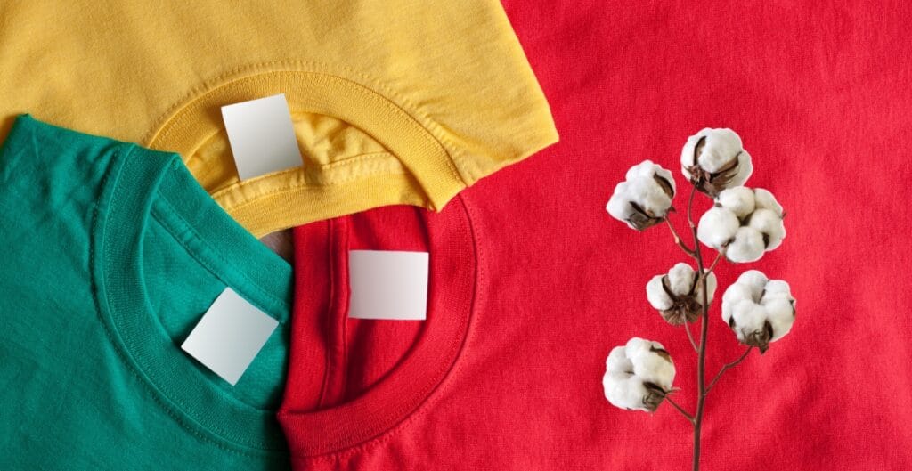 Three colorful T-shirts (yellow, green, and red) are folded and stacked with blank white tags, placed next to a cotton branch on a red fabric background.