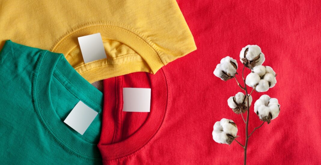 Three colorful T-shirts (yellow, green, and red) are folded and stacked with blank white tags, placed next to a cotton branch on a red fabric background.