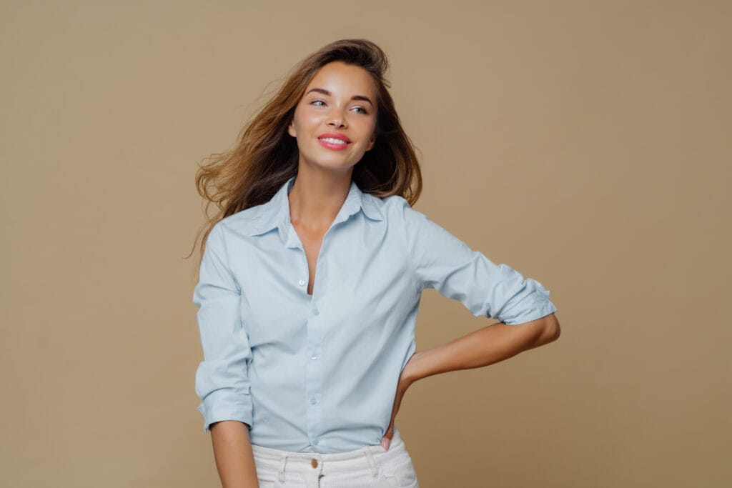 A woman with long, wavy brown hair wearing a light blue chambray shirt, smiling softly and posing with one hand on her hip against a beige background.