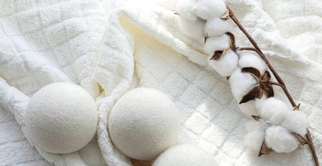 A soft, white, textured fabric background with two round, white bath bombs, and a cotton branch with fluffy white cotton bolls.