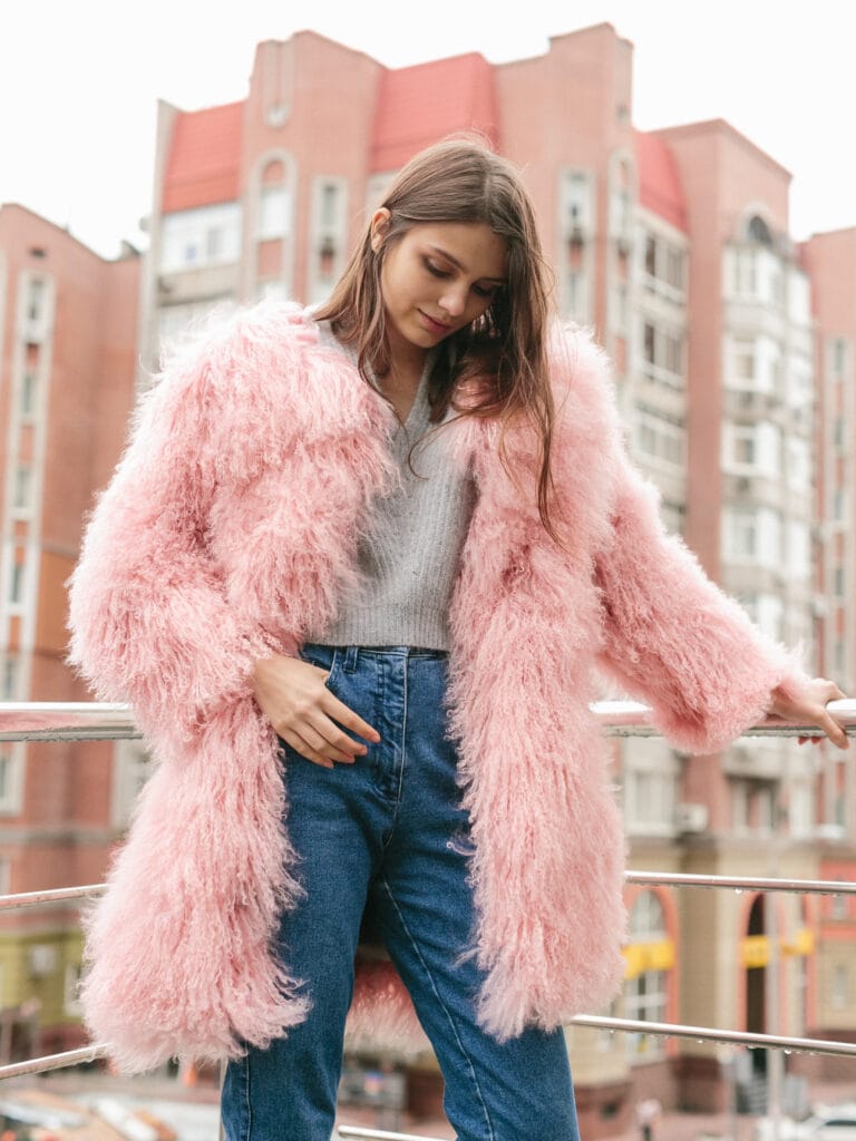A young woman wearing a fluffy pink fuzzy coat, standing on a balcony with a cityscape of brick buildings behind her.