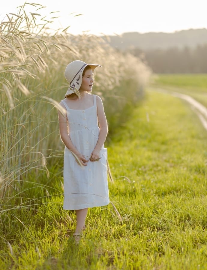 A young girl in a white pinafore cotton dress standing in a grassy field with tall grasses, wearing a wide-brimmed hat, and gazing into the distance on a sunny spring day.