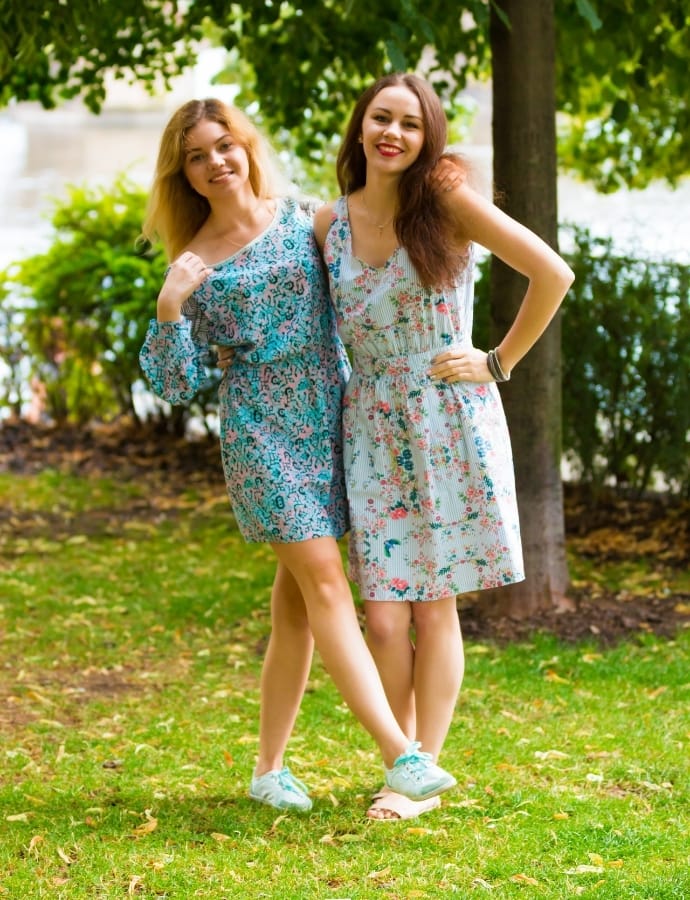 Two women standing outdoors in a park, wearing colorful summer cotton pinafore dresses with floral and abstract patterns, smiling and enjoying the sunny day.