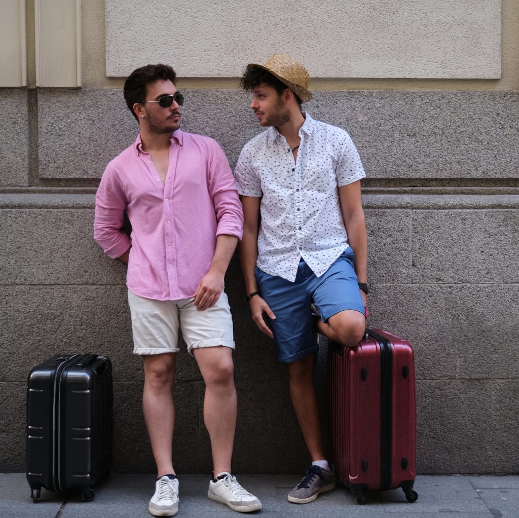 Two stylish young men standing against a textured wall, each with a suitcase. The man on the left is wearing sunglasses, a pink short-sleeve button-down shirt, beige shorts, and white sneakers. The man on the right is wearing a straw hat, a white short-sleeve button-down shirt with a small pattern, blue shorts, and grey sneakers, with one foot resting on his pink suitcase.