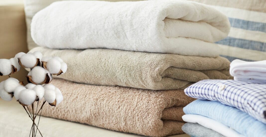 A neatly stacked pile of towels in neutral tones, with a bunch of cotton stems on the side, placed on a light-colored surface.