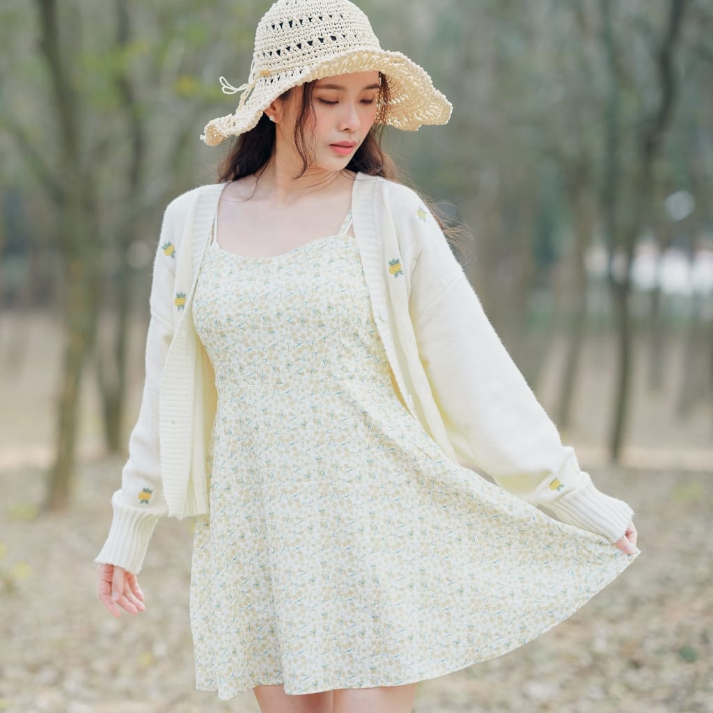A young woman wearing a light-colored floral dress, a white cardigan with little embroidered details, and a wide-brimmed straw hat, standing outdoors among trees. She is gently holding the hem of her dress, with a soft and peaceful expression.
