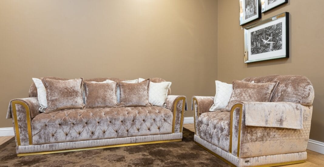 Two elegant sofas upholstered in plush velvet fabric, with the larger sofa featuring a button-tufted design, multiple cushions, and a luxe texture, placed against a neutral wall with framed black-and-white photographs.