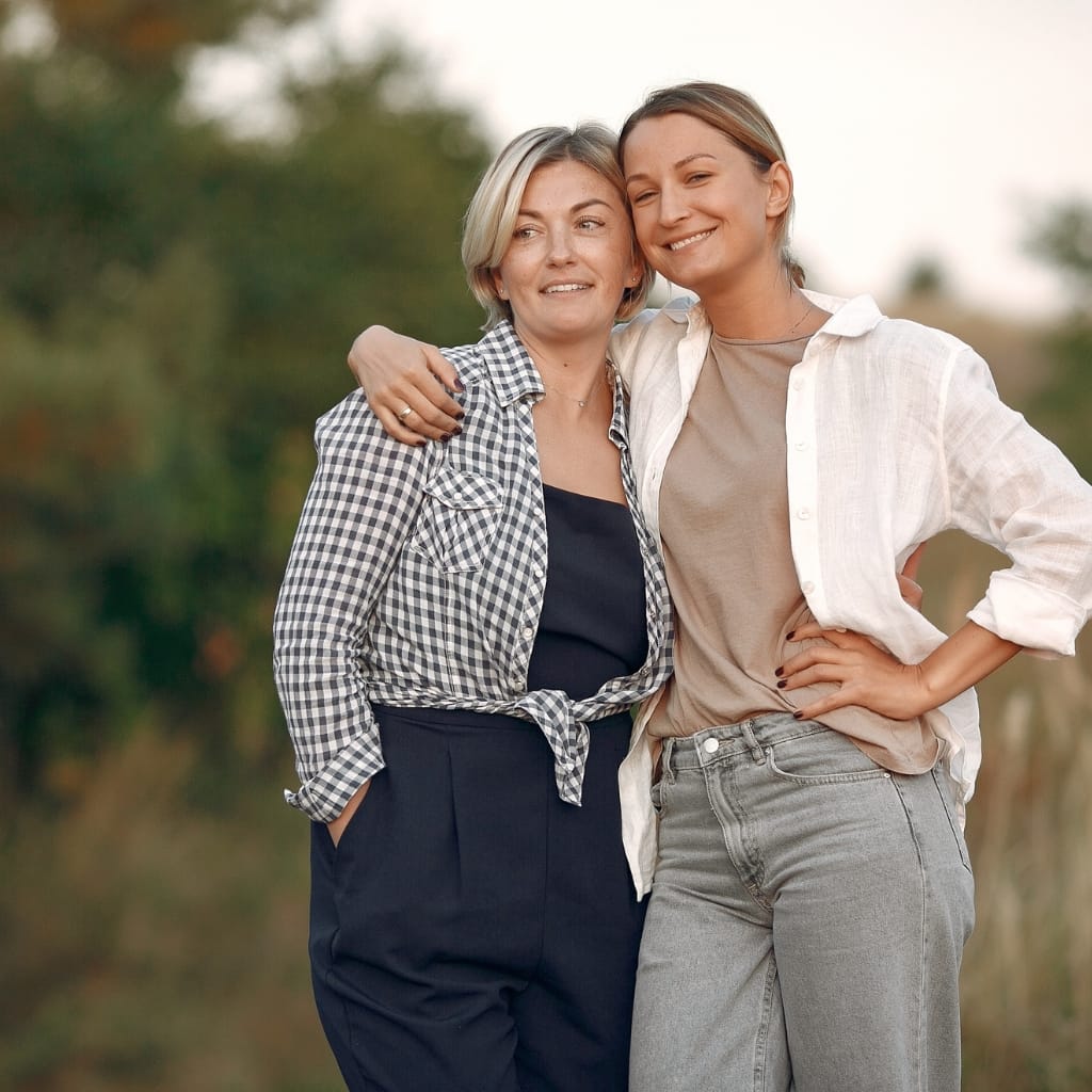 Two women, likely from different generations, stand close together outdoors. The woman on the right has her arm around the shoulder of the woman on the left, both smiling warmly.