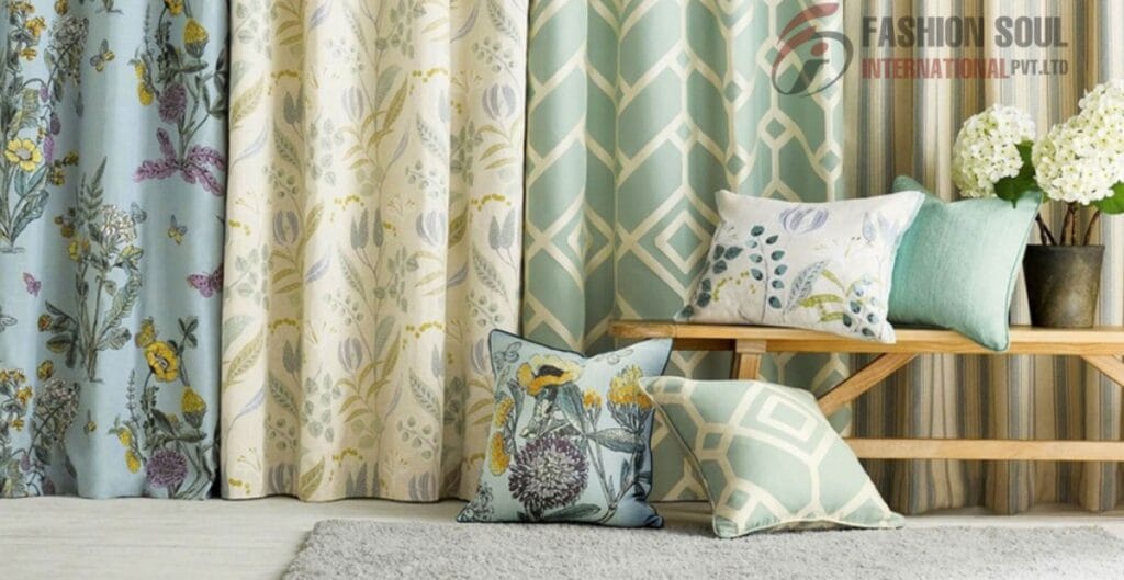 A cozy interior scene featuring a variety of patterned curtains and decorative pillows. The curtains have floral and geometric designs in pastel shades of blue, cream, and yellow. The pillows, also patterned, include floral and geometric motifs, arranged on a light-colored surface near a wooden bench and a vase with white flowers.