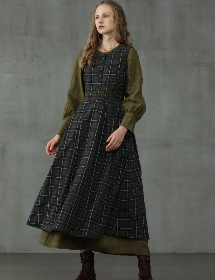A young woman standing against a concrete wall, wearing a layered winter outfit featuring a dark plaid pinafore dress over a long-sleeve olive green top, paired with dark boots.
