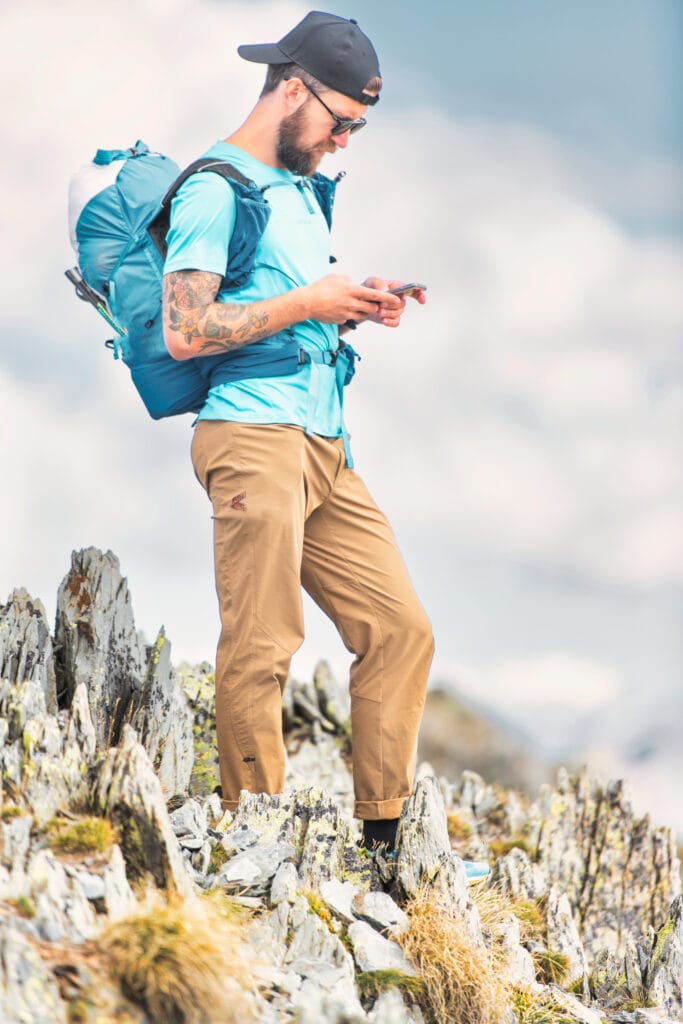 A man wearing outdoor hiking attire, including a blue shirt, tan pants, and a backpack, standing on rocky terrain and looking at his phone.