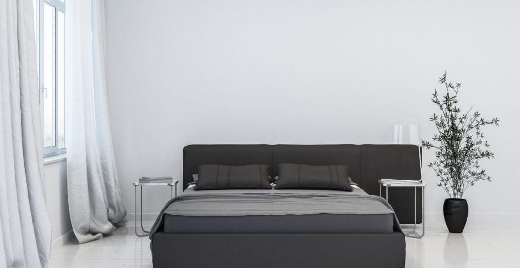 A modern bedroom with black linen bedding, featuring a neatly made bed with two matching black pillows, positioned against a white wall. There are two small side tables with books, a large black vase with a tall green plant, and white curtains on a window to the left.