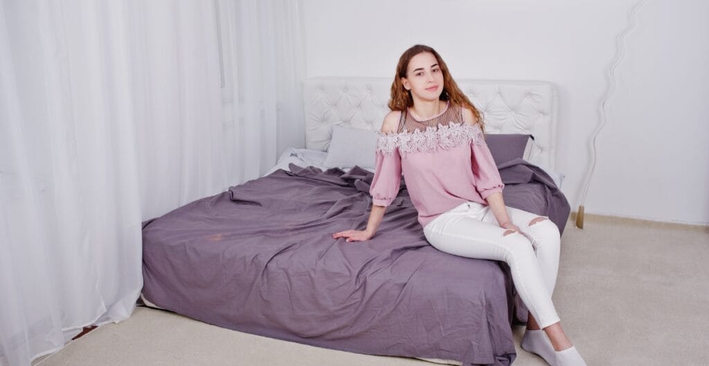 A young woman with long wavy hair, wearing a pink blouse and white pants, sitting on the edge of a bed in a bright bedroom with white walls and curtains.