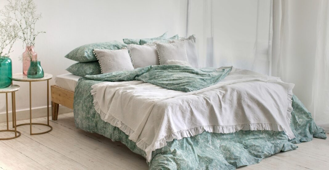 A neatly made bed with green and white bedding, including multiple pillows, in a bright room with white curtains and wooden flooring. Two small side tables with vases and decorative plants are beside the bed.