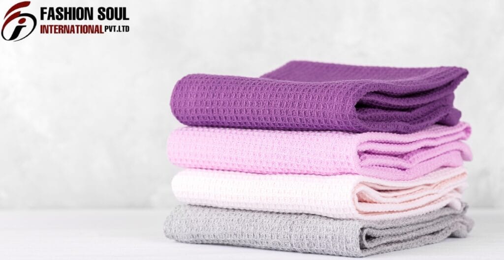 A stack of four folded waffle-knit towels in purple, pink, white, and gray, placed on a white surface against a light background with a "FASHION SOUL INTERNATIONAL PVT. LTD" logo in the top-left corner.