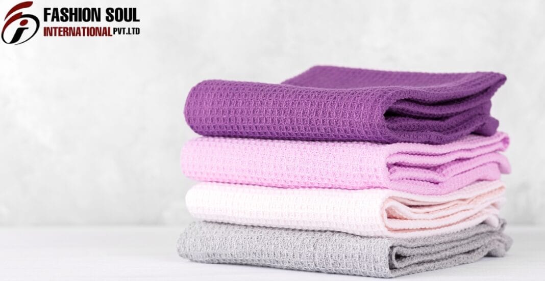 A stack of four folded waffle-knit towels in purple, pink, white, and gray, placed on a white surface against a light background with a 