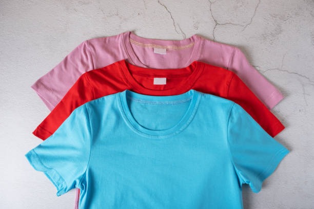 Three plain short-sleeve t-shirts in blue, red, and pink colors stacked on top of each other on a light textured surface.