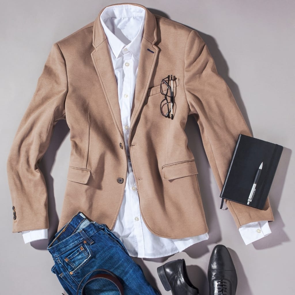 A professional men's outfit laid out on a flat surface, including a tan blazer with a white shirt underneath, a pair of blue jeans with a brown belt, black dress shoes, a black notebook with a pen placed on top, and a pair of eyeglasses hanging from the blazer pocket.
