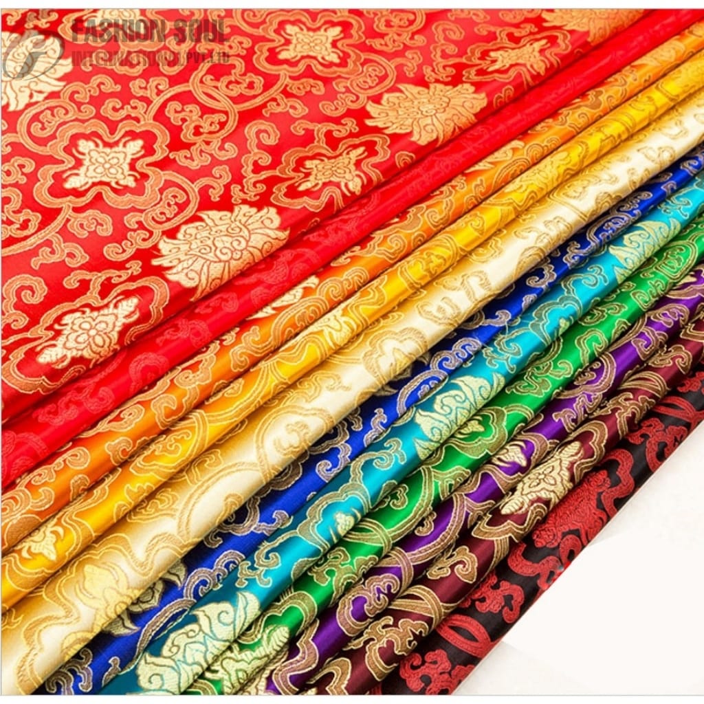 A collection of colorful, intricately patterned silk fabrics arranged in a diagonal row. The fabrics feature traditional floral and swirling designs in various vibrant colors, including red, orange, yellow, blue, green, purple, and black, all accented with gold detailing.