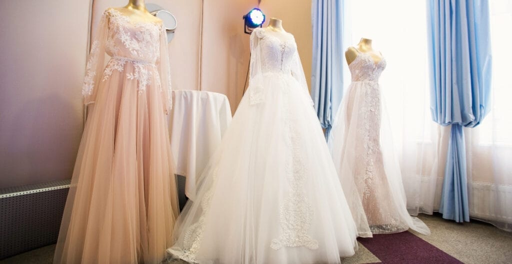 Four elegant ball gowns displayed on mannequins, each with intricate lace and tulle details, set against a softly lit background with blue curtains.