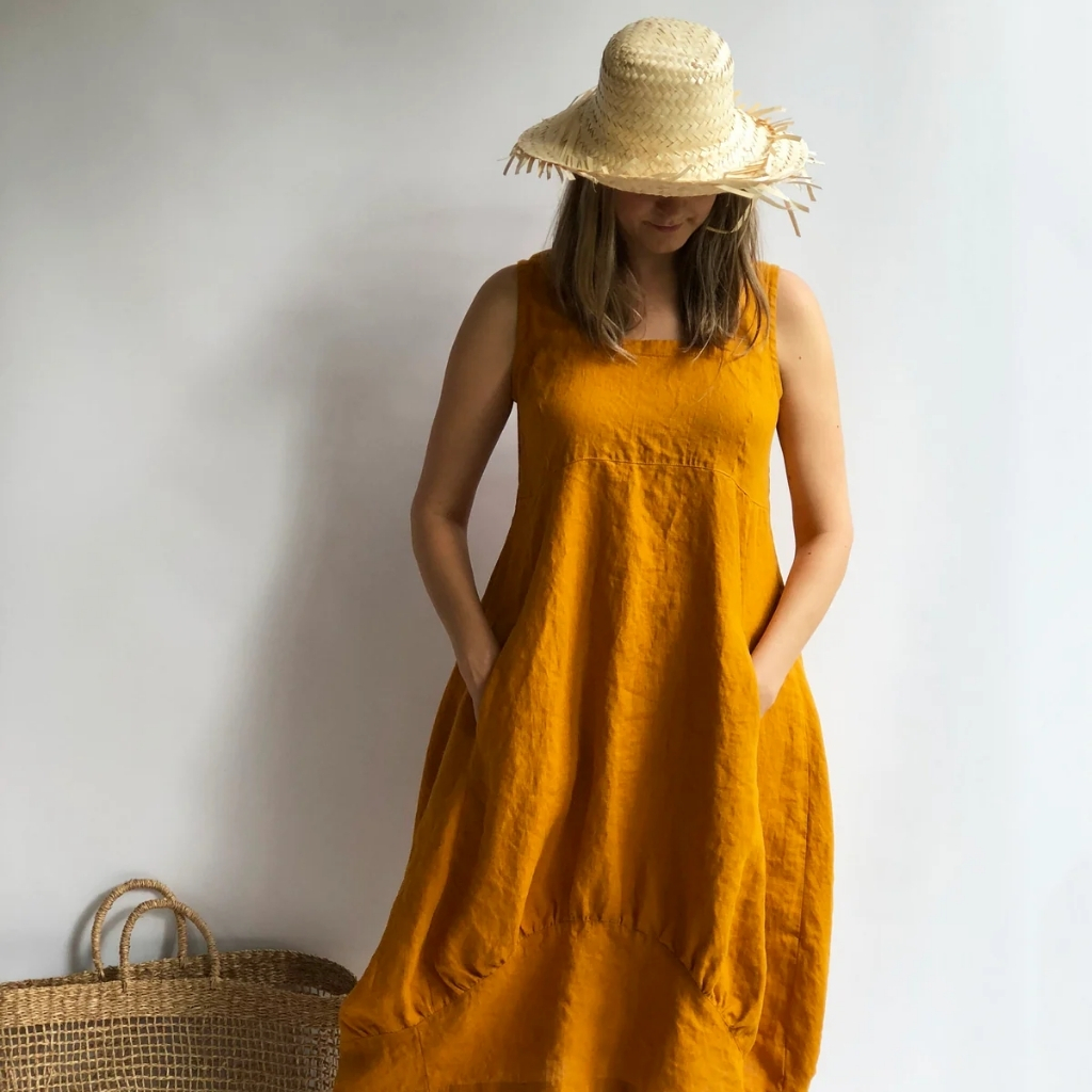 A woman wearing a wide-brimmed straw hat and an orange sleeveless dress, standing against a plain white background with her hands in her pockets.