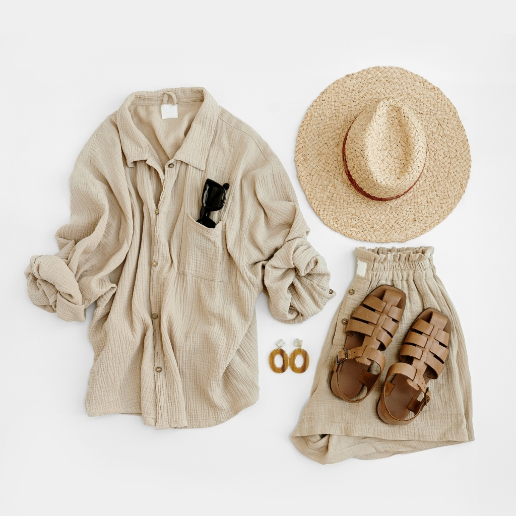  Flat lay of a casual summer outfit with a beige oversized shirt, a wide-brimmed straw hat, a pair of brown woven sandals, a pair of sunglasses in the shirt pocket, gold earrings, and a beige skirt, all arranged on a white background.